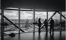 Three people stand by large windows, gazing out at a panoramic view of the city skyline.